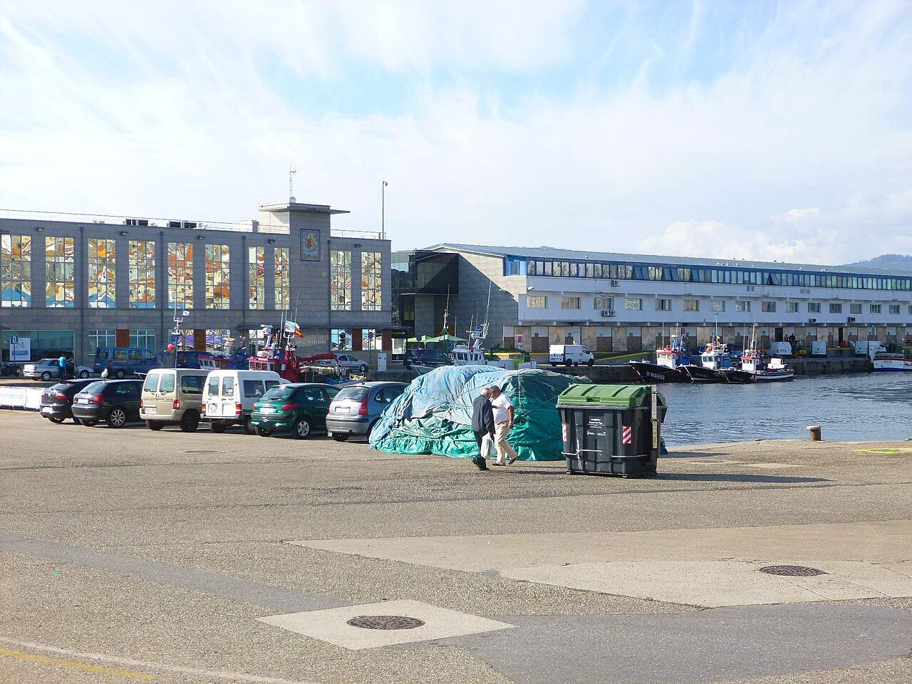 Muelle del puerto pesquero de Vigo con coches aparcados, aparejos cubiertos con lonas, edificios portuarios y varios barcos pesqueros amarrados al fondo.
