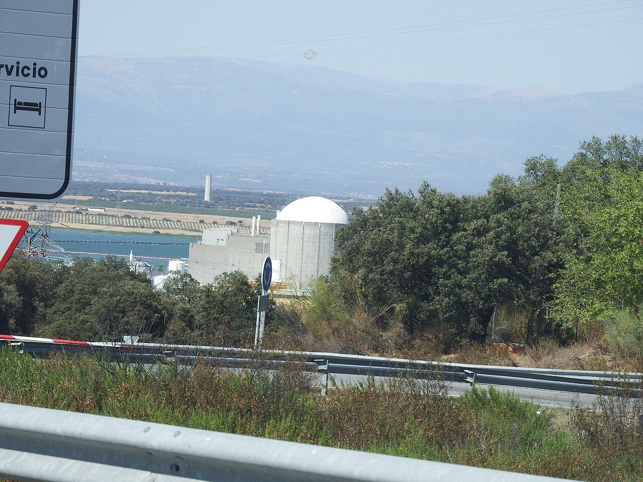 Edificio de un reactor con cúpula blanca junto a un embalse, visto desde una carretera con una señal de tráfico en primer plano