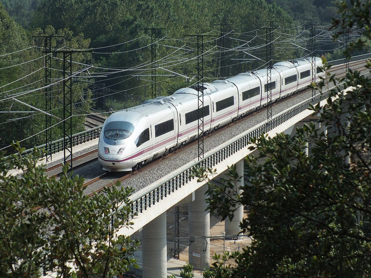 Tren de alta velocidad blanco con franja morada sobre un viaducto de hormigón con catenaria, entre árboles