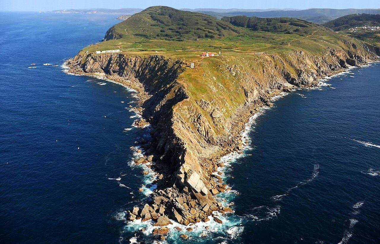 Vista aérea de un cabo rocoso alargado que entra en el mar, con un faro y algunos edificios en la punta