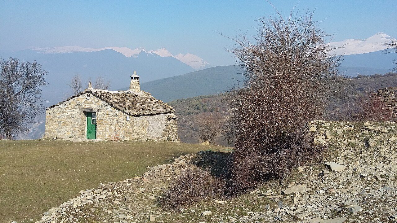 Casa de piedra con tejado de losas junto a muros derruidos y matorral, con montañas nevadas del Pirineo al fondo.