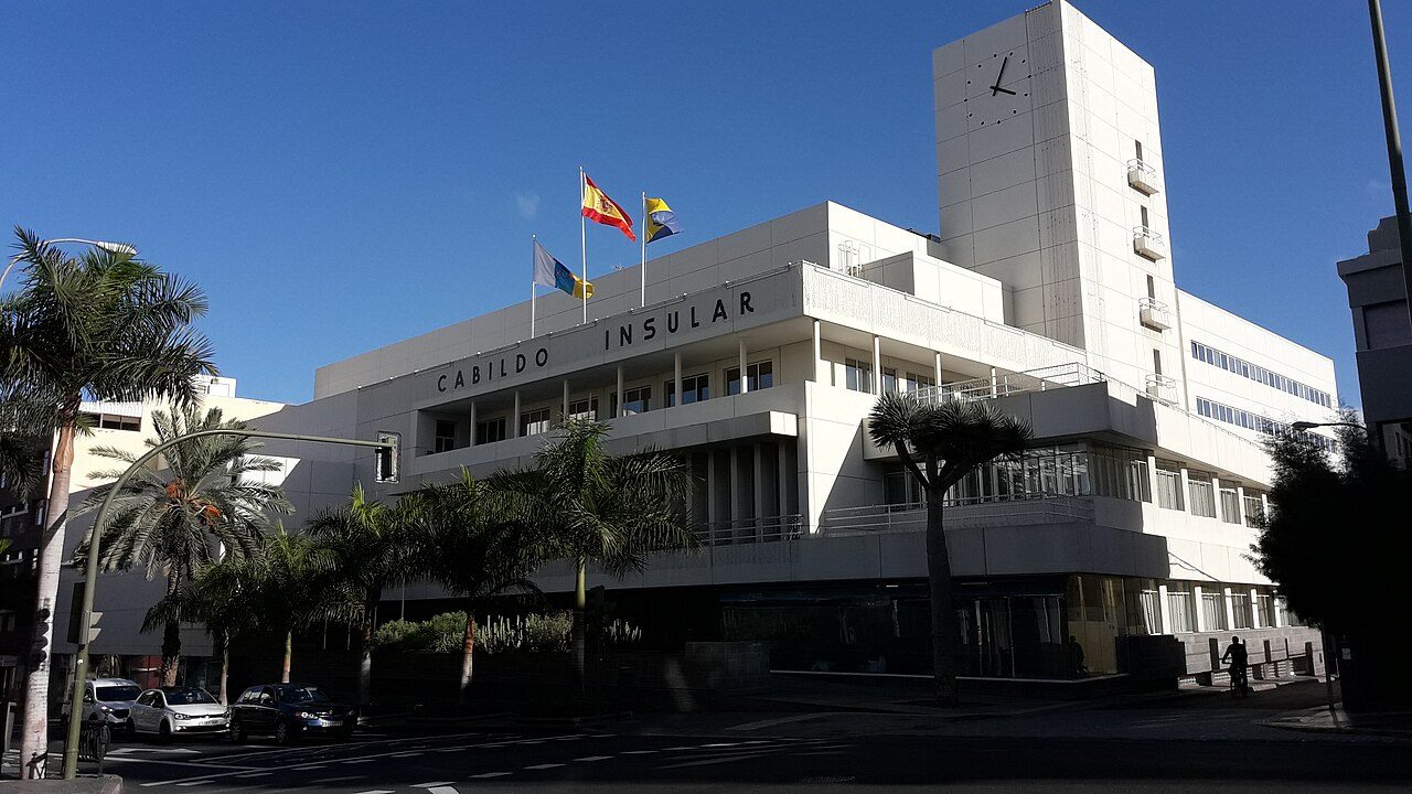 Edificio blanco de líneas rectas, con torre de reloj, banderas y el rótulo «Cabildo Insular», entre palmeras.