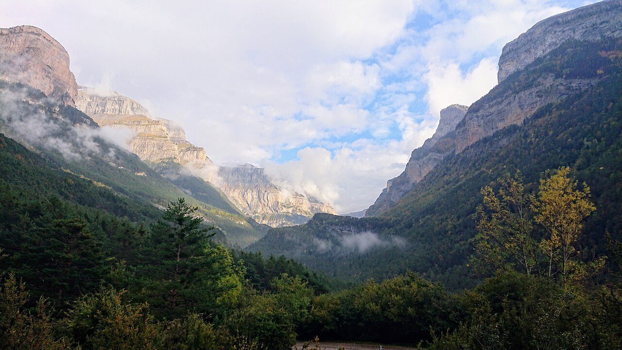 Valle ancho en forma de U con las laderas cubiertas de bosque y grandes paredes verticales de roca caliza en lo alto, entre nubes