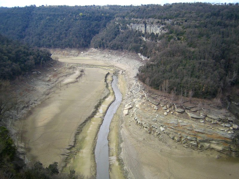 Embalse casi vacío, con el antiguo cauce del río serpenteando por un fondo de barro y laderas de bosque a los lados.