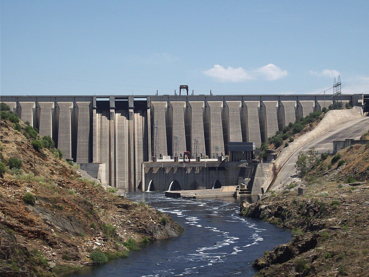 Gran presa de hormigón de contrafuertes vista desde aguas abajo, con el río saliendo por la base en un valle encajado