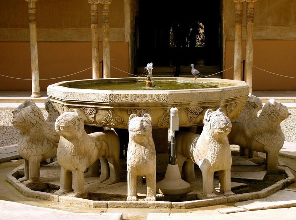 Fuente de piedra con una taza sostenida por doce leones y, al fondo, columnas finas de una galería