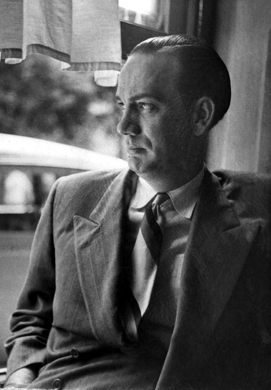Fotografía en blanco y negro de Camilo José Cela joven, con traje y corbata, sentado junto a una ventana
