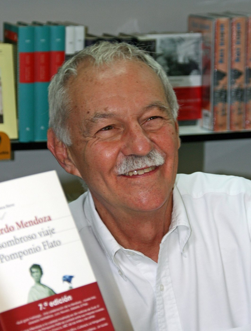 Eduardo Mendoza, sonriente, con canas y bigote, enseña un ejemplar de su novela El asombroso viaje de Pomponio Flato en la Feria del Libro